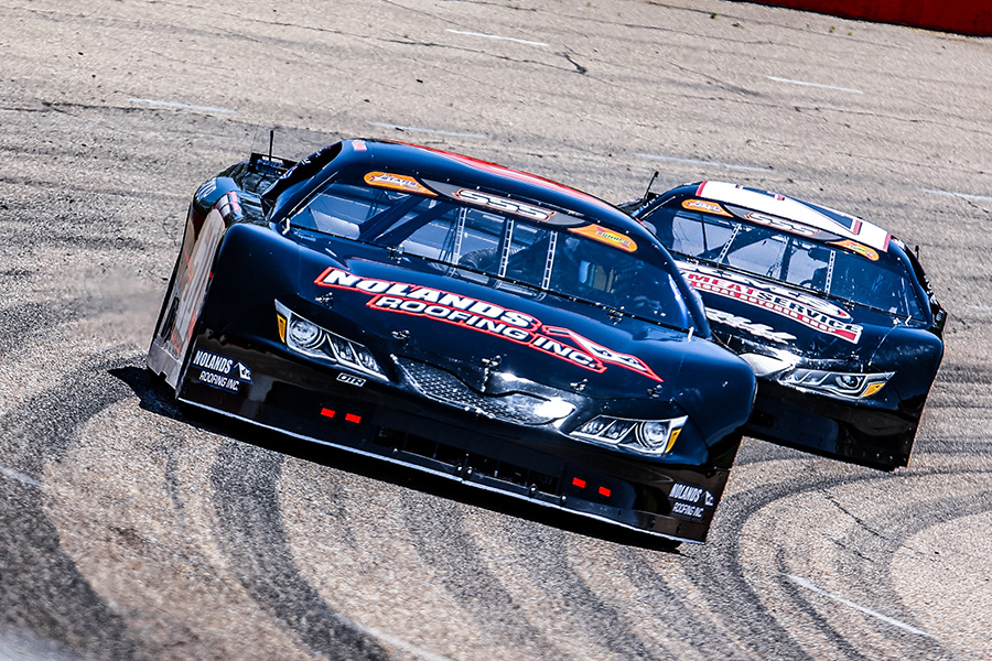 Two black stock cars, one prominently displaying 'Noland's Roofing Inc.' on the front, race closely on a sunlit track. The leading car is closely followed by another, with both cars navigating a turn, leaving tire marks on the asphalt.