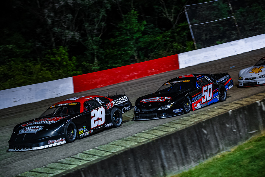 Super Late model cars, number 29 and number 50(Jett Noland), race side by side on a dimly lit track during a night event, with a third car trailing behind. The cars navigate a curve with a red and white barrier, surrounded by dense trees.
