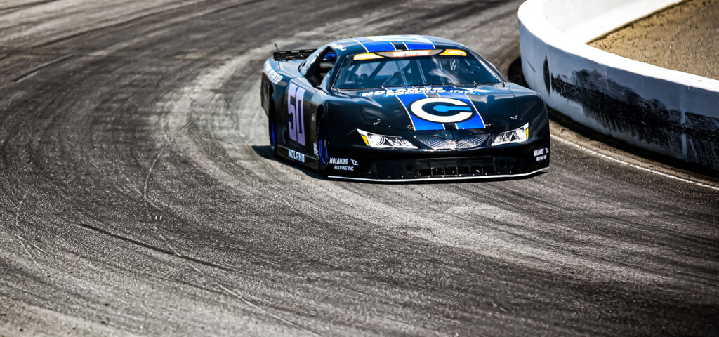 Jett Noland driving the #50 Pro Late Model car during the Pro Late Model 100 race, hugging the curve of the racetrack in a sleek black and blue livery.